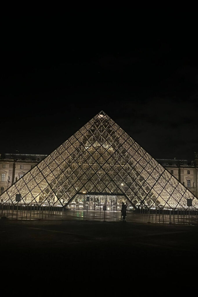 The Louvre Pyramid spectacularly lit at night, casting a warm glow and standing as a contrast to the dark evening sky, with the silhouette of a person in the foreground giving scale to the grandeur of the scene.