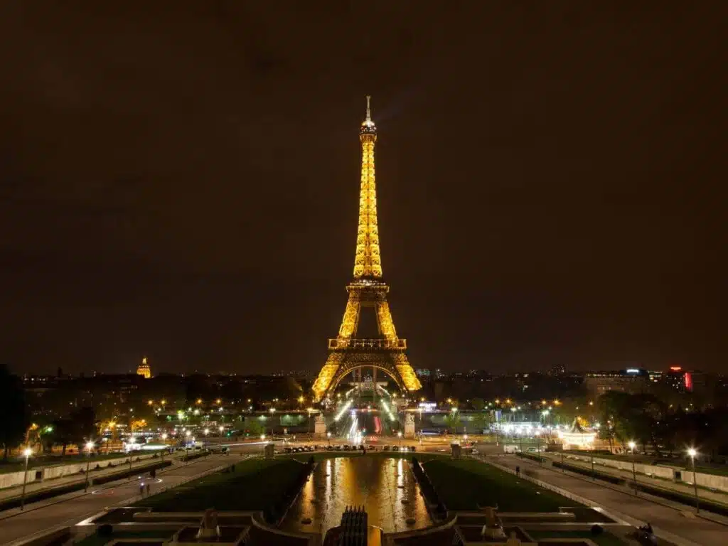 Night view of the Eiffel Tower illuminated with golden lights, with the cityscape and dark sky in the background. The surrounding area is lit with streetlights and the Seine River is faintly visible, reflecting some lights.