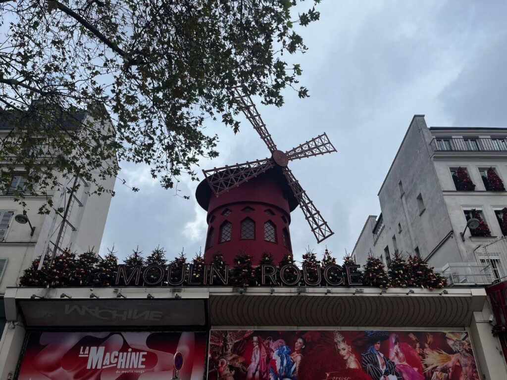 he iconic red windmill atop the Moulin Rouge in Paris, adorned with festive decorations, against a cloudy sky with leafy branches framing the view.