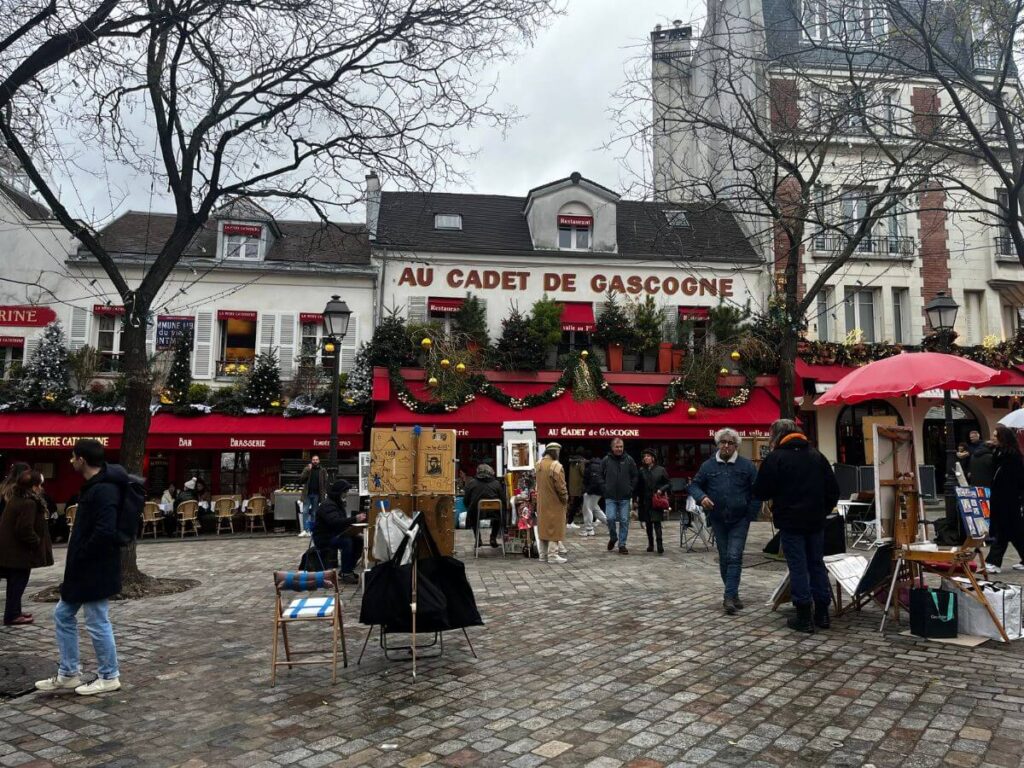 Busy square in Paris with people browsing through market stalls, artists displaying their work, and the decorated facade of 'Au Cadet de Gascogne' restaurant.
