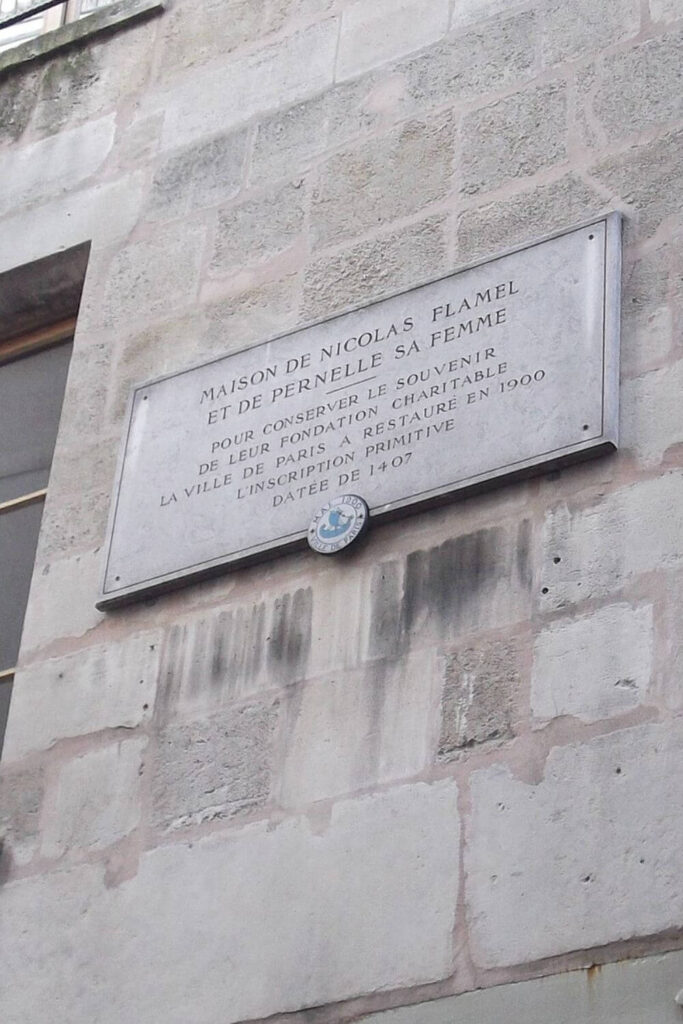 A commemorative plaque on a weathered stone wall, inscribed with 'Maison de Nicolas Flamel et de Pernelle sa femme' in French, marking the historical significance of the site related to Nicolas Flamel and his wife Pernelle.