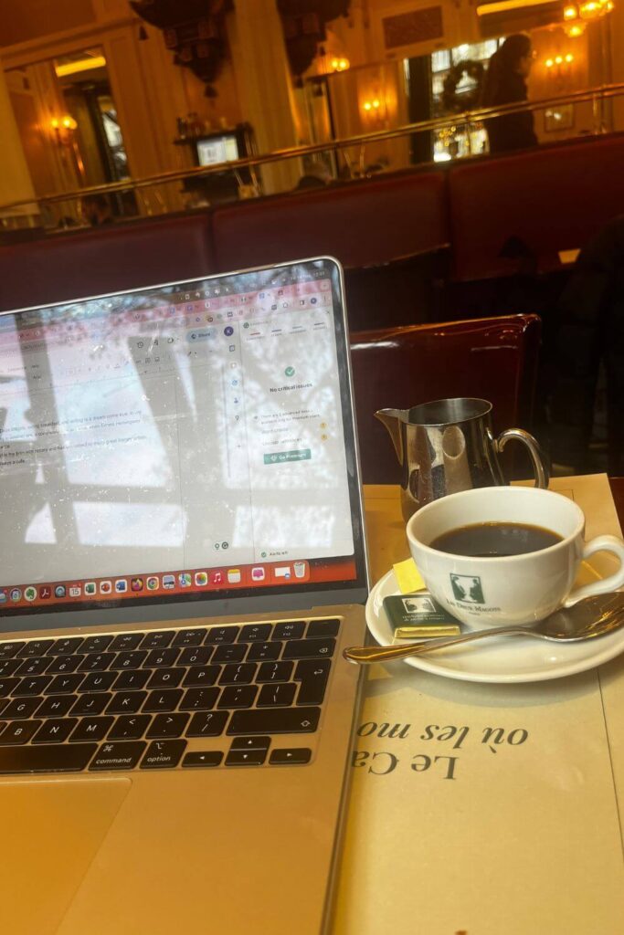A writer's morning at Les Deux Magots café in Paris, with a laptop open on a wooden table, accompanied by a classic cup of black coffee and a silver pot, reflecting the ambient interior lighting of the café.