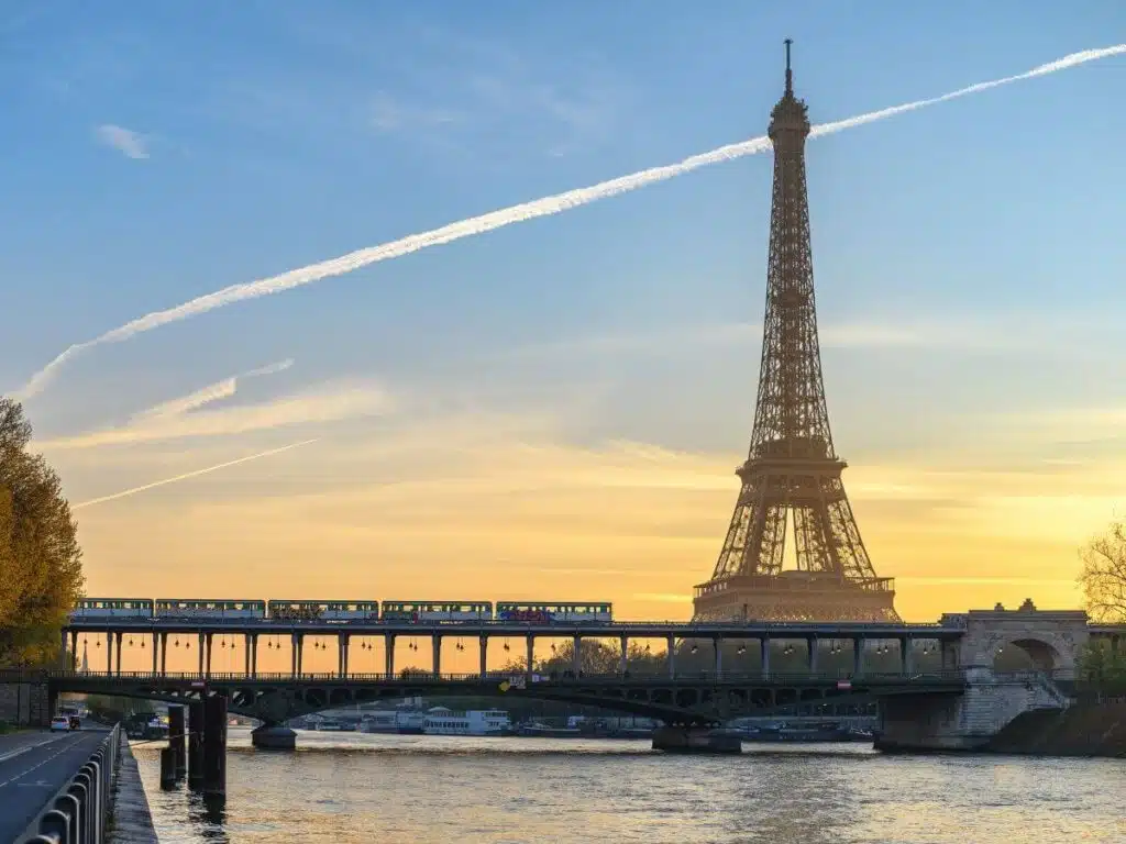 Sunset view of the Eiffel Tower with the Seine River in the foreground. A metro train crosses the Pont de Bir-Hakeim bridge, while the sky above is adorned with streaks of airplane contrails. The warm glow of the setting sun bathes the scene in soft light.