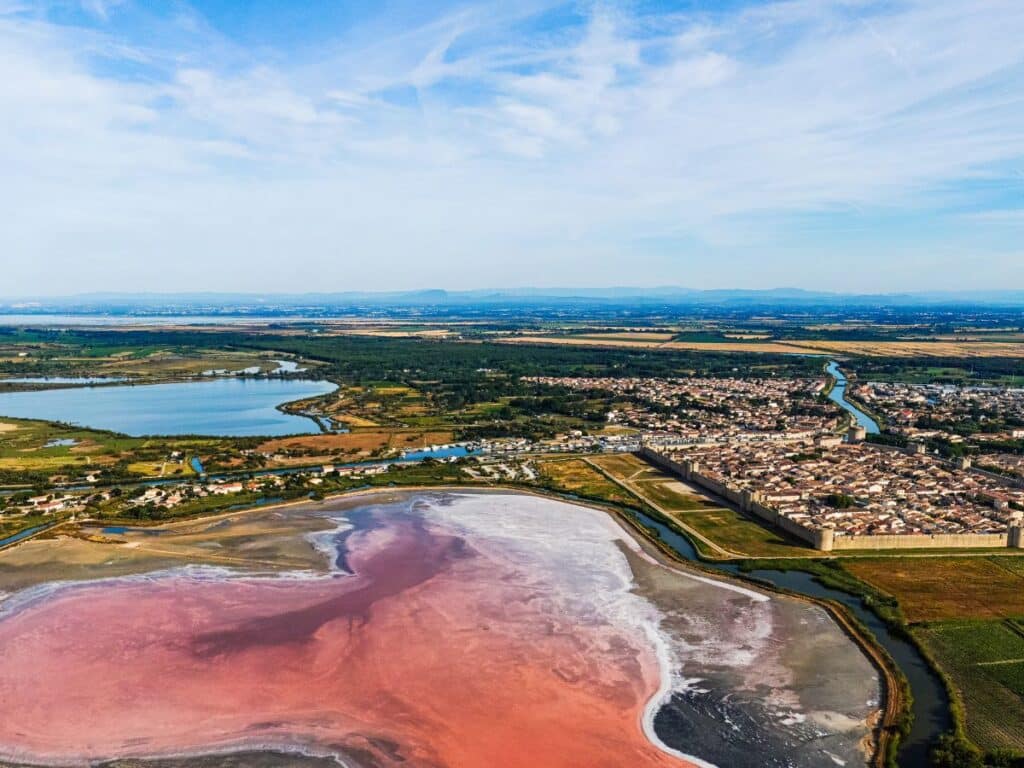 Aerial view of the pink salt flats of Aigues-Mortes in Occitanie, with the fortified town and meandering waterways in the background, set against a vast landscape leading to the horizon.