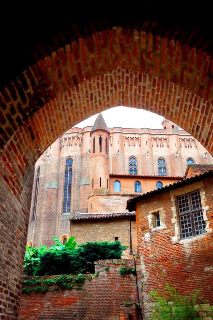 View of the towering brick facade of Albi Cathedral, seen through a semicircular archway, contrasting the weathered textures of the surrounding buildings in Albi, Midi-Pyrénées, with lush greenery adorning the walls.