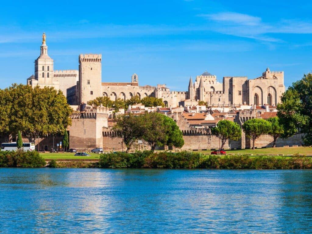 The historic Papal Palace in Avignon, Provence-Alpes-Côte d’Azur, viewed from across the Rhône River, with its imposing fortified walls and towers against a clear blue sky.