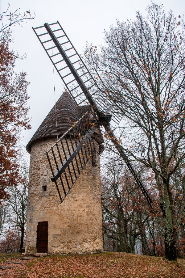 Traditional stone windmill with wooden blades in Beaumont-du-Périgord, set against a backdrop of bare trees and a cloudy winter sky.