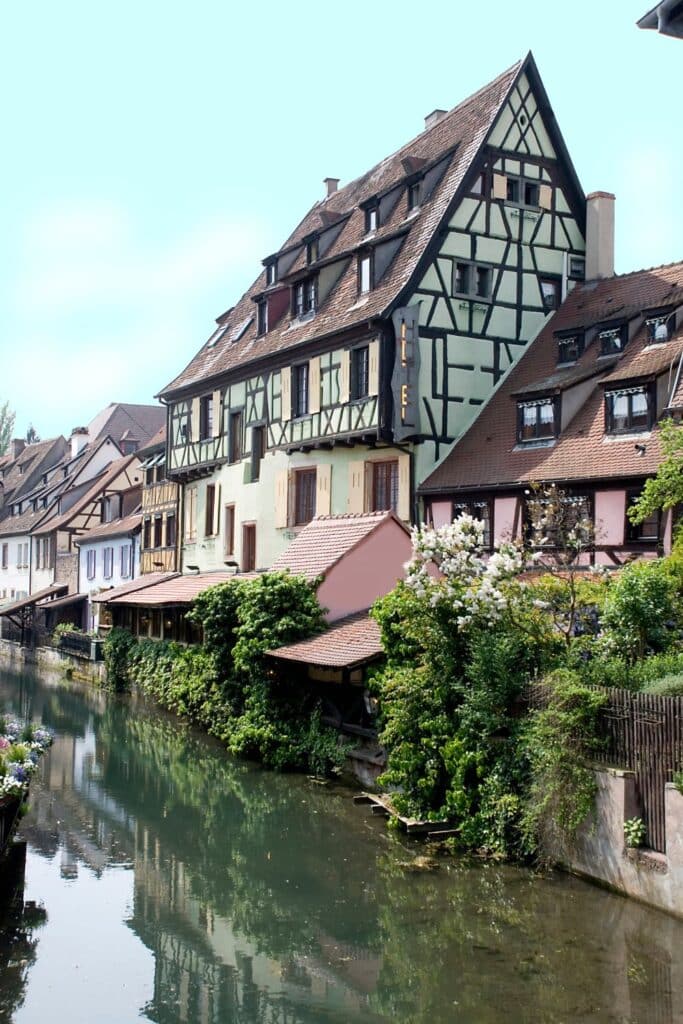 Half-timbered houses with traditional Alsatian architecture reflected in the calm waters of a canal in Colmar, France, on a bright day, showcasing the charming and picturesque 'Little Venice' area of the town.