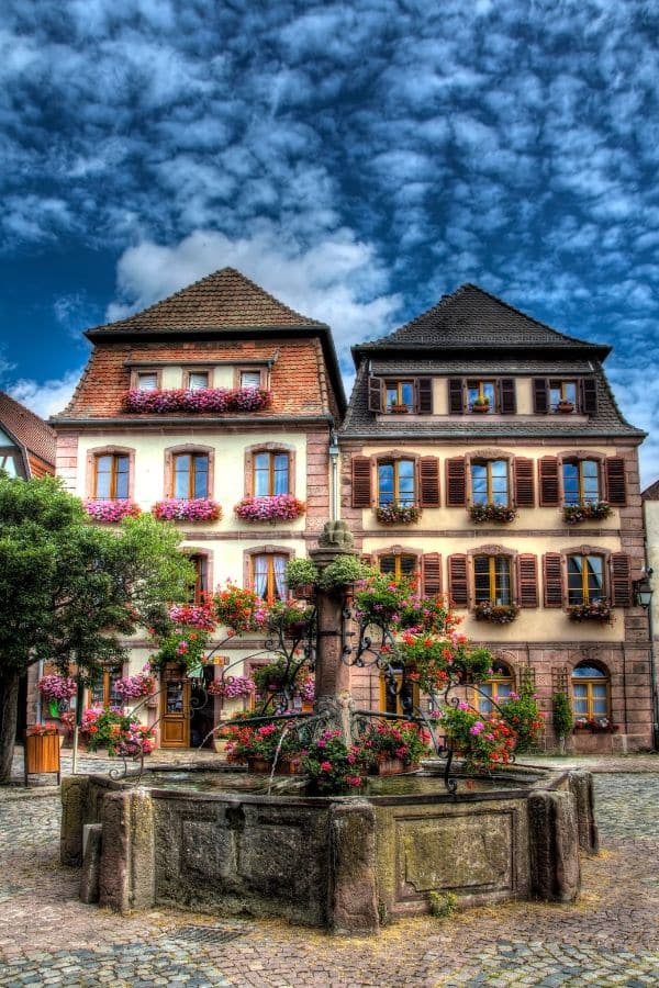 Two tall houses side by side with three window going across each level with shutter and window boxes with pink flowers. A water fountain with greenery stands in front of them