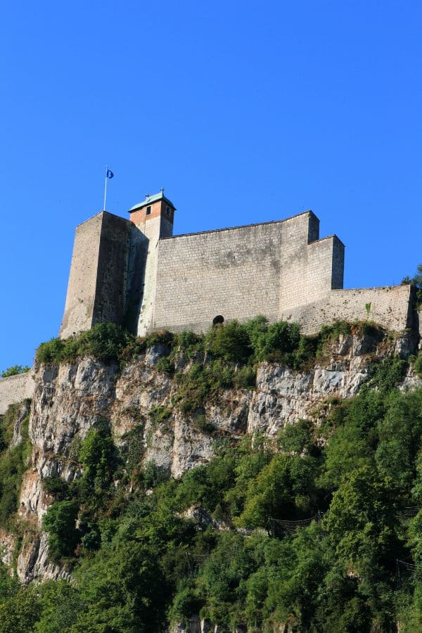Imposing citadel of Besançon, perched on a rugged cliff, dominating the landscape against a clear blue sky, with lush green foliage accentuating the ancient fortification's stone walls.