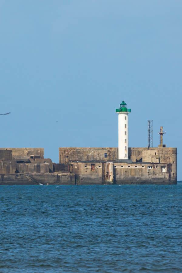 Close-up view of the white lighthouse with a green top at the harbor of Boulogne-sur-Mer, against a blue sky with a hint of clouds, and the sea in the foreground.