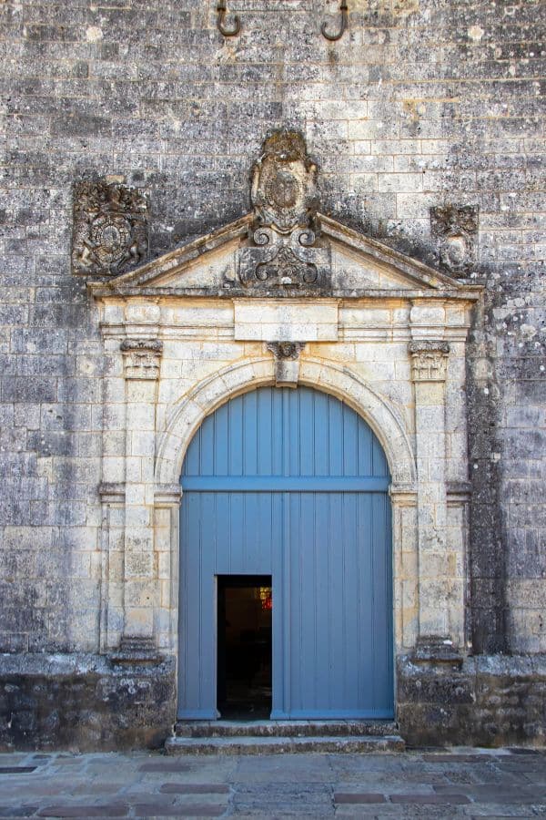 Ornate stone doorway of the historical fortress in Brouage, Charente-Maritime, featuring a classical pediment with a coat of arms and weathered masonry, contrasted by a bright blue wooden door slightly ajar.