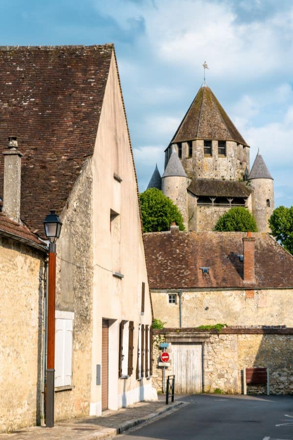 The medieval Cesar Tower in Provins, France, rises majestically behind rustic limestone buildings, with a streetlamp in the foreground, set against a serene blue sky with soft clouds.