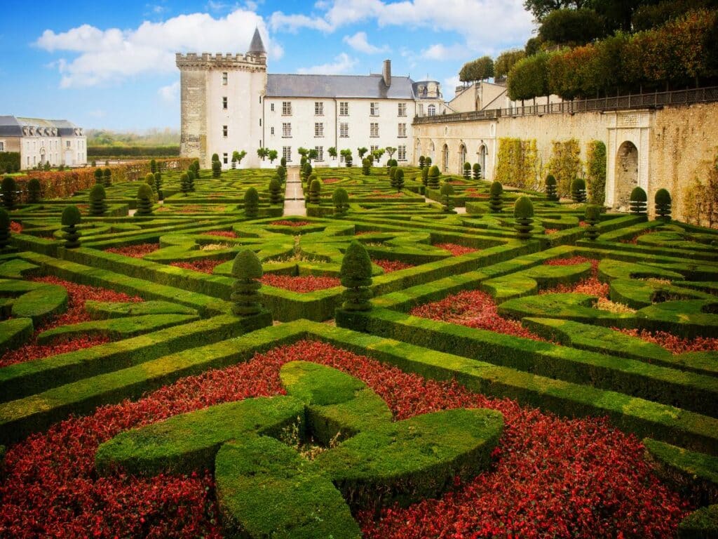 Ornate geometric garden of Chateau de Villandry, Loire Valley, with intricate patterns of green hedges and vibrant red flowers, set against the historic castle and its stone walls under a partly cloudy sky.