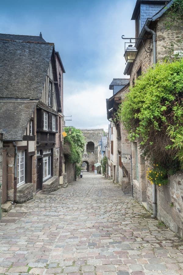 Cobbled street in Dinan, Brittany, lined with traditional half-timbered houses and overflowing greenery, leading towards an archway in the distance, under a soft cloudy sky.