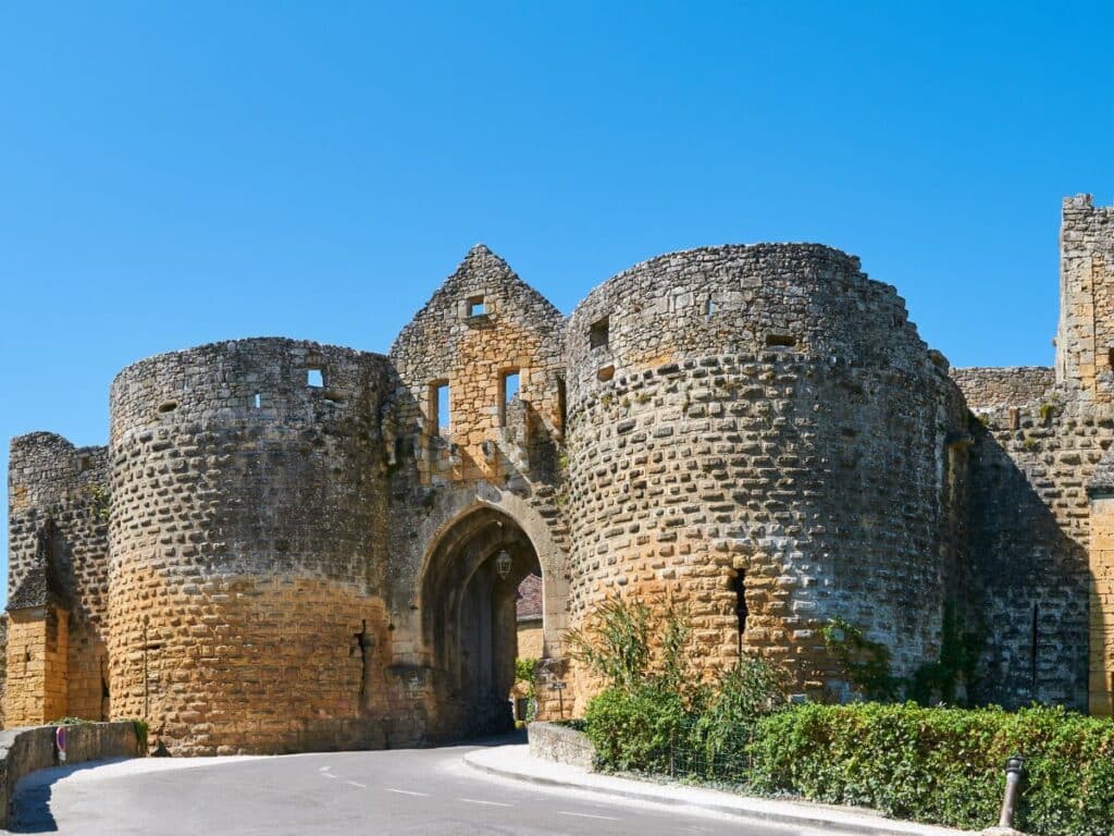 Ancient stone gatehouse of Domme, Dordogne, with its arched entrance and remnants of towers, standing resolute under a clear blue sky.