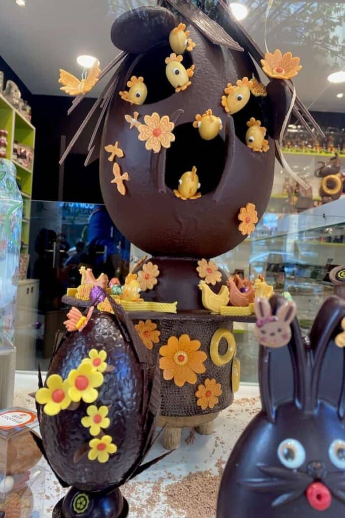 Intricately crafted dark chocolate Easter egg adorned with small yellow chick decorations and orange flowers, showcasing French artisan chocolate traditions.