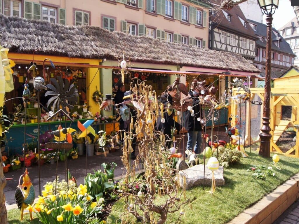 A vibrant outdoor Easter market in France, featuring colorful decorations such as pinwheels, hanging ornaments, metal sculptures, and bright yellow daffodils, set against charming half-timbered buildings.