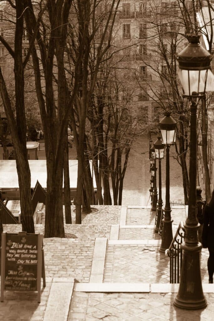 Sepia-toned image of a descending staircase in Montmartre, Paris, lined with vintage street lamps and bare trees, with a chalkboard sign displaying a quote: 'The Irish have a very simple taste, they are always satisfied with the best - Oscar Wilde.