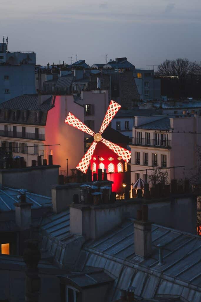An illuminated red windmill, the Moulin Rouge, with white lattice patterns stands out against the twilight sky, surrounded by the rooftops of Parisian buildings. The scene captures a distinctive landmark, glowing vibrantly in the evening.