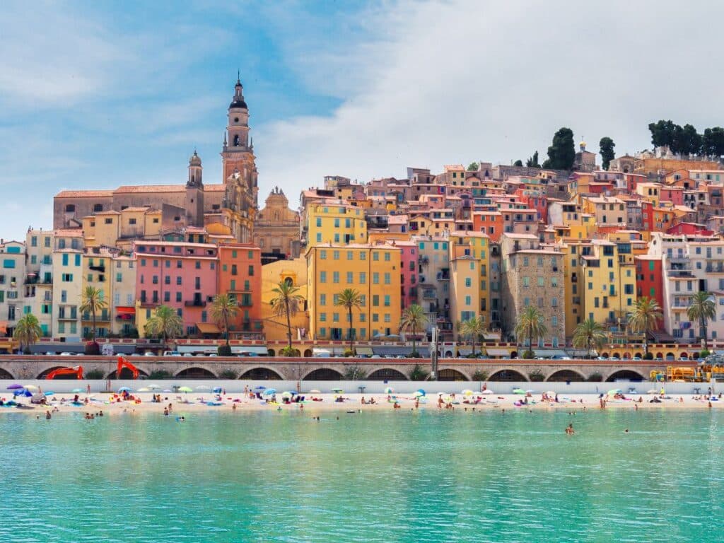 A serene beachfront in Menton, France, with azure waters and sunbathers enjoying the pebbled shore. The town's historic architecture, painted in warm pastel hues, rises in the background, crowned by the stately basilica and bell tower. Palm trees line the promenade, completing this picturesque Riviera setting, inviting relaxation and exploration.