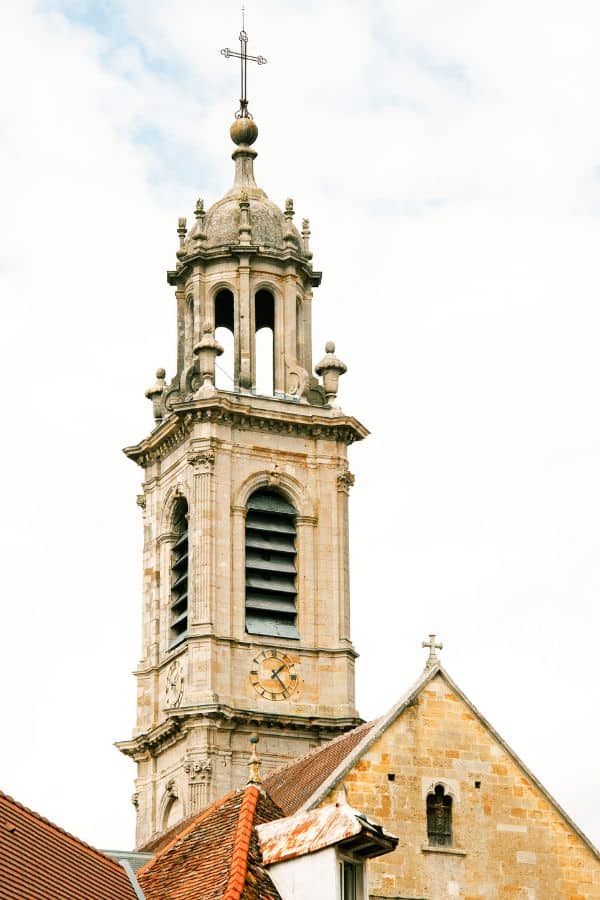 The baroque bell tower of the Cathedral of Saint-Mammès in Langres, Haute-Marne, with its ornate stonework and clock face, stands tall above surrounding rooftops against a cloudy sky.