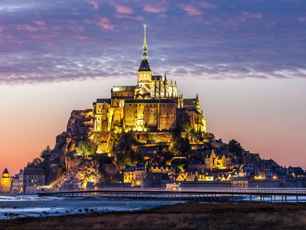 Twilight view of Mont Saint-Michel in Normandy, illuminated against the dusk sky, with its Gothic architecture and spire standing out above the surrounding bay and causeway.