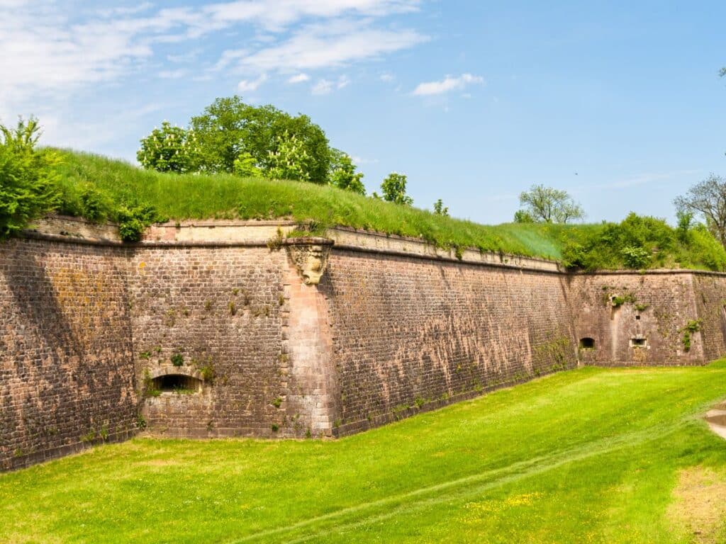 Well-preserved fortification walls of Neuf-Brisach in Alsace, with lush green grass at the base and atop the walls, under a bright blue sky with soft white clouds.