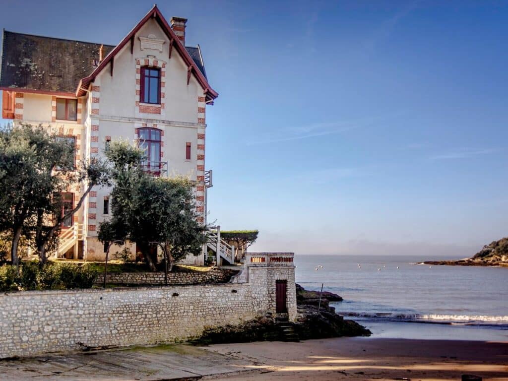 "Charming traditional house with red and white façade in Pontaillac, Royan, nestled among green trees with a view of the calm sea and a clear sky at dawn.