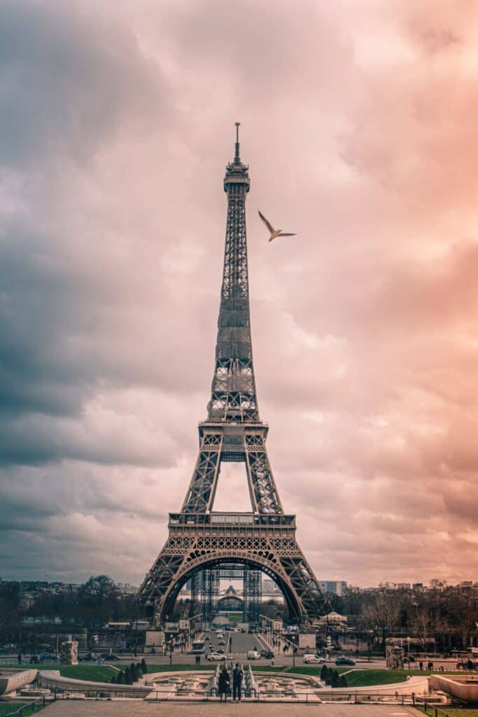 Dramatic view of the Eiffel Tower against a cloudy sky with warm tones, as seen from the Champ de Mars gardens, with a single bird flying high near the top and people visible in the foreground, capturing the iconic majesty of Paris.