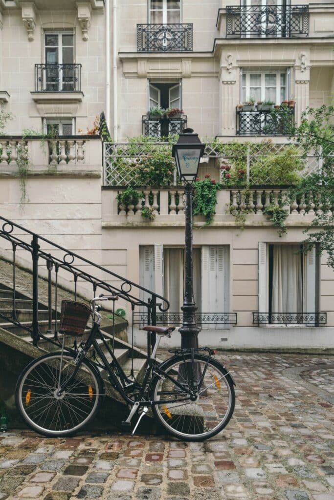 A quaint Parisian courtyard scene with a black bicycle leaning against a wrought iron railing beside stone steps, with a traditional street lamp and lush balcony gardens in the background, evoking a serene, everyday moment in France.