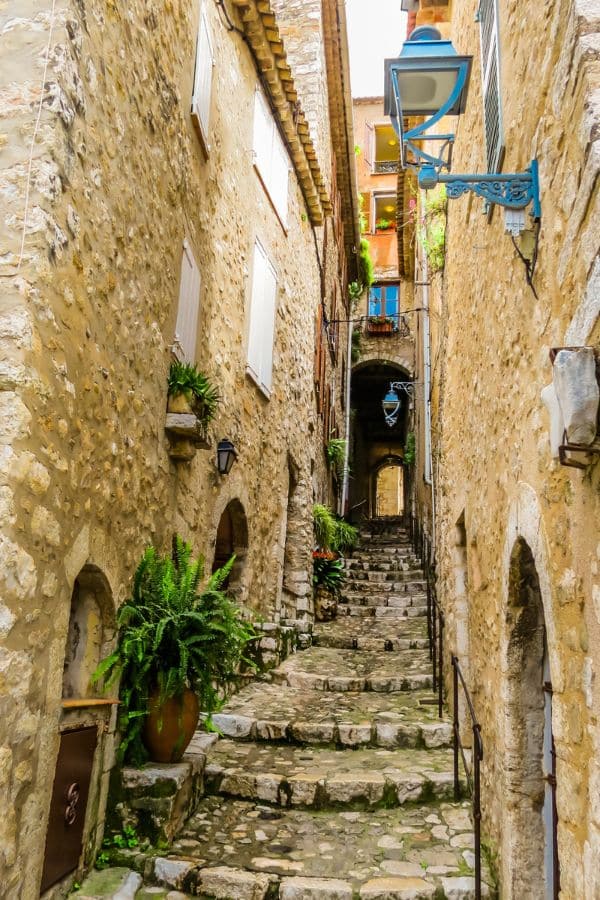 Narrow, ascending cobblestone alleyway in Saint-Paul de Vence, lined with historic stone buildings adorned with green plants and hanging lanterns, evoking a sense of quaint medieval charm.