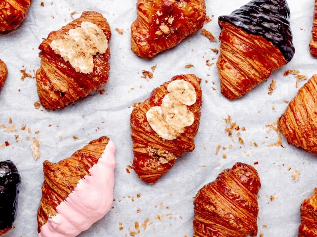 An assortment of gourmet croissants with various toppings such as sliced banana, pink frosting, nuts, and chocolate glaze, arranged on parchment paper.