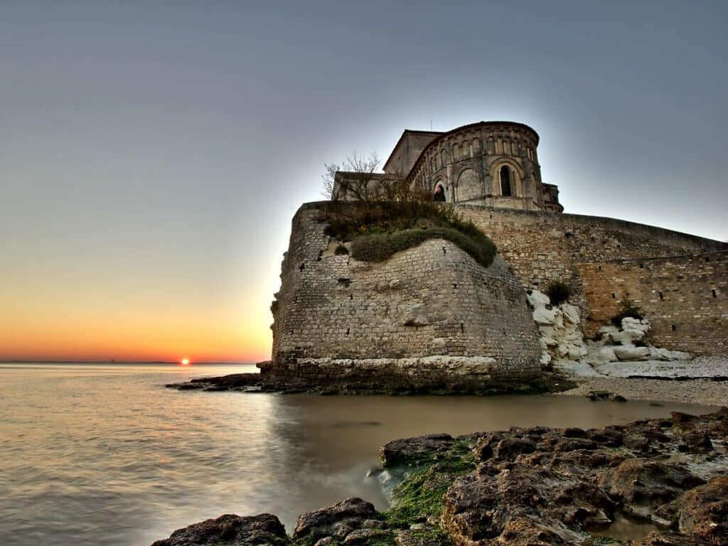 Sunset view at Talmont-sur-Gironde, Charente-Maritime, featuring the historic church perched on a fortified cliff overlooking the sea, with the sun dipping below the horizon.