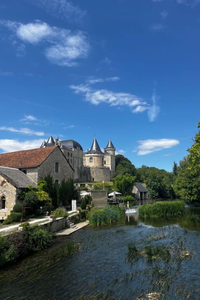 Calm waters and lush riverside foliage frame the imposing Chateau of Verteuil, its majestic towers and stone walls standing as a testament to centuries-old French architecture. Wisps of cloud in the bright blue sky complement the serene and dignified presence of this historic landmark in the heart of the French countryside.