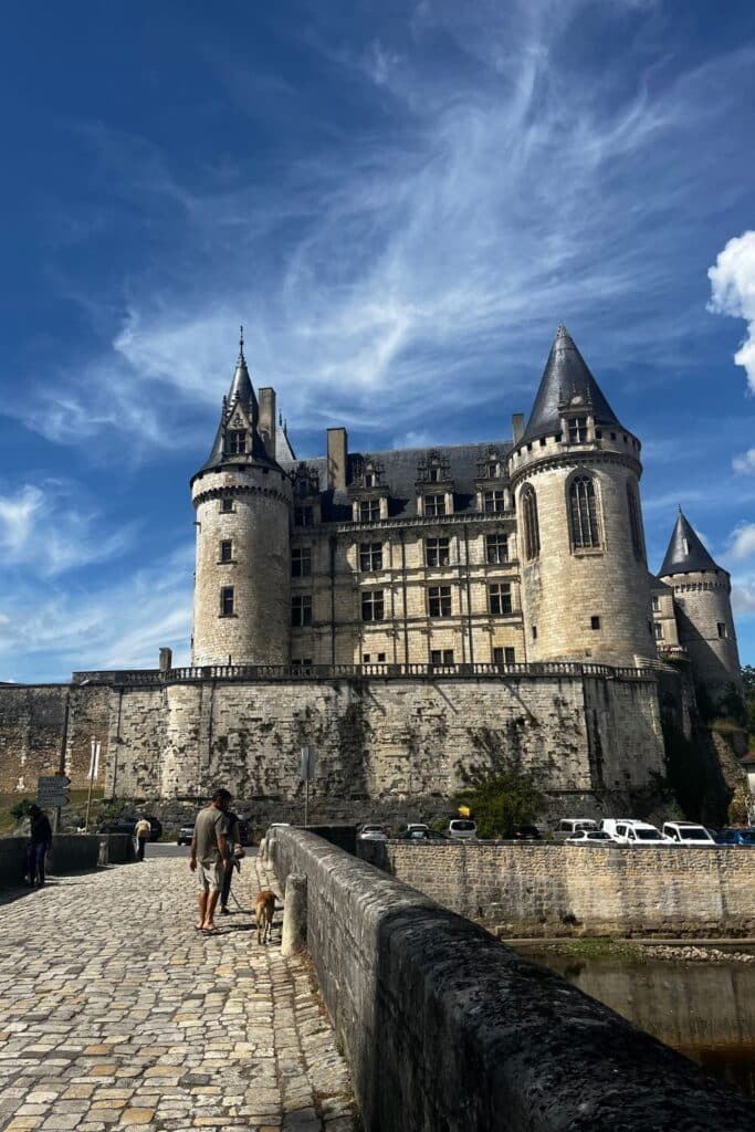 A person walks their dog along the stone bridge leading to the imposing Château de La Rochefoucauld, its grand towers and fortified walls standing against a backdrop of blue sky and wispy clouds. This historic French castle, steeped in the legacy of the Rochefoucauld family, dominates the landscape, a striking example of Renaissance architecture.