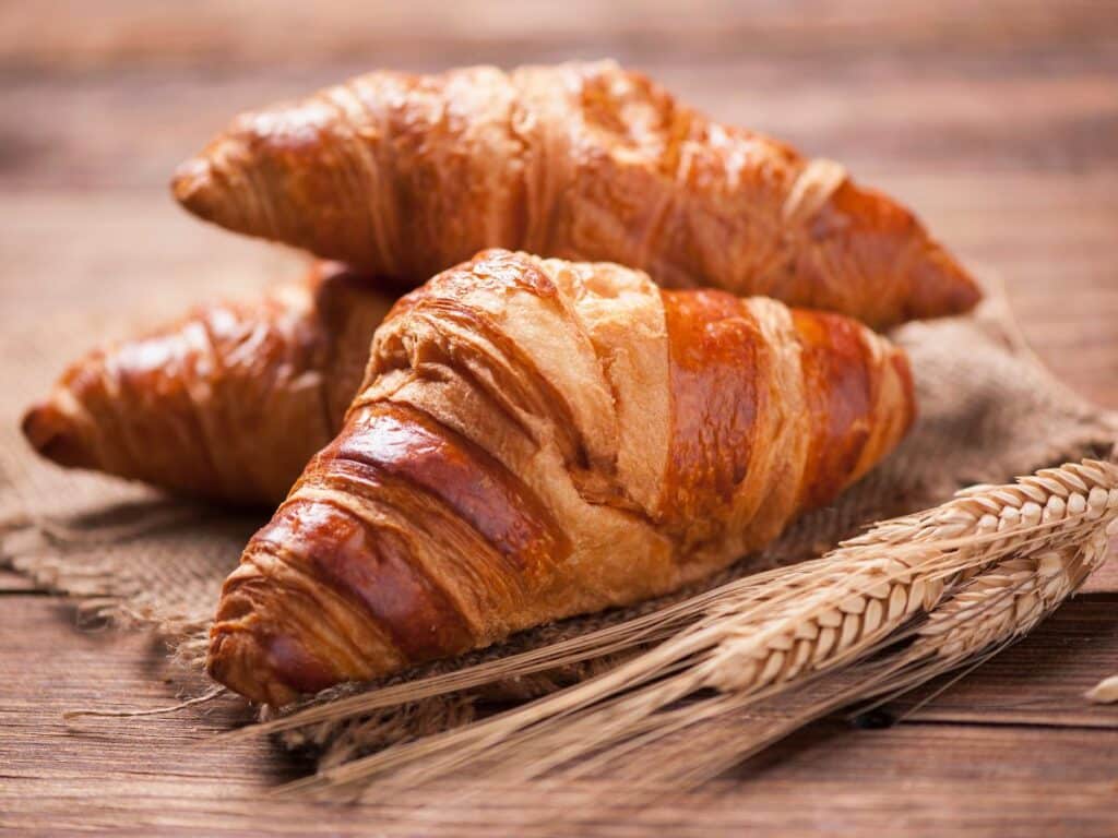 Freshly baked croissants with a golden crust, displayed on a wooden table alongside wheat stalks, symbolizing National Croissant Day in France.