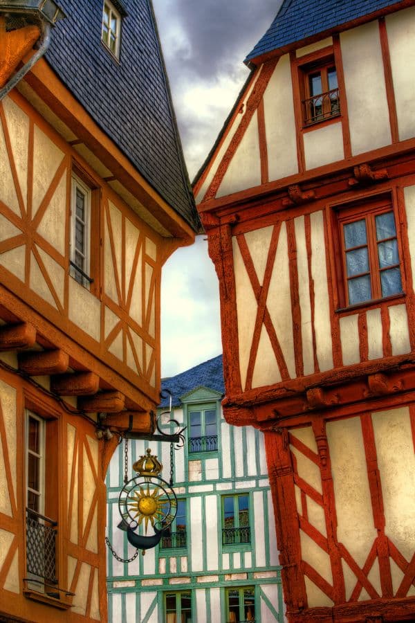 Vividly detailed close-up of traditional half-timbered houses in Vannes, Brittany, with intricate wood framing and a decorative hanging lantern, set against an overcast sky.