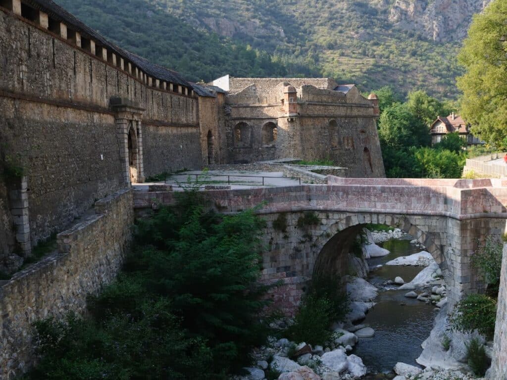 Ancient stone bridge and fortified walls of Villefranche-de-Conflent, nestled in a lush valley with a gentle river flowing underneath, framed by the greenery and mountains of rural France.