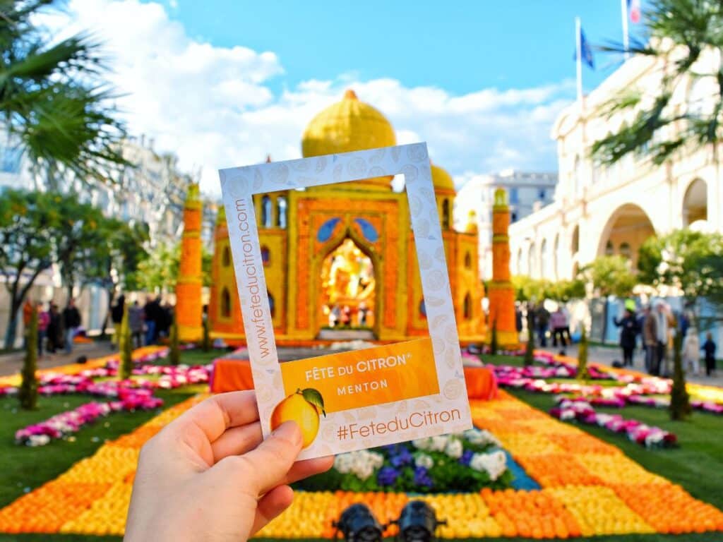 A hand holds up a pamphlet with the "Fête du Citron® Menton #FeteduCitron" and the event's website against a backdrop of elaborate citrus sculptures and floral arrangements. The festival's vibrant displays and attendees can be seen under a clear blue sky, hinting at the festive and colorful atmosphere of the Menton Lemon Festival.