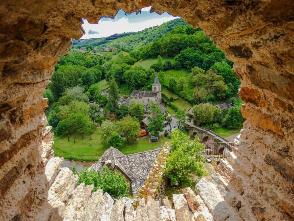 Rustic village view through a stone window, with a church, bridge, and lush trees.