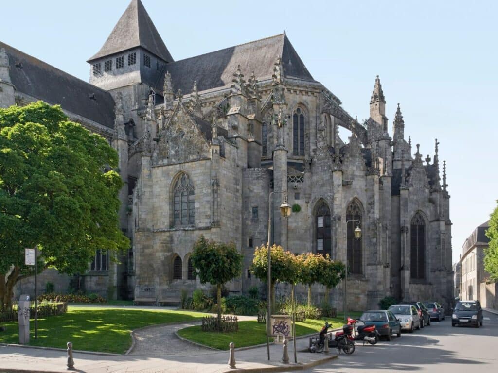 Basilique Saint-Sauveur de Dinan, a majestic Gothic and Romanesque church, stands proudly amidst greenery and modern street life on a sunny day, highlighting a blend of historical reverence and contemporary urban life.