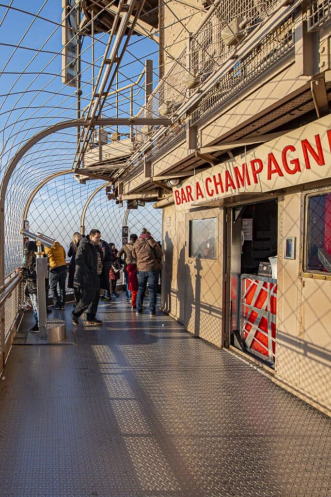 Visitors gather at the 'Bar à Champagne' on the Eiffel Tower's observation deck, enjoying the view behind a safety net, with the Parisian cityscape below them. The late afternoon sun casts long shadows across the metal grid floor, adding to the ambiance of this unique high-altitude experience.