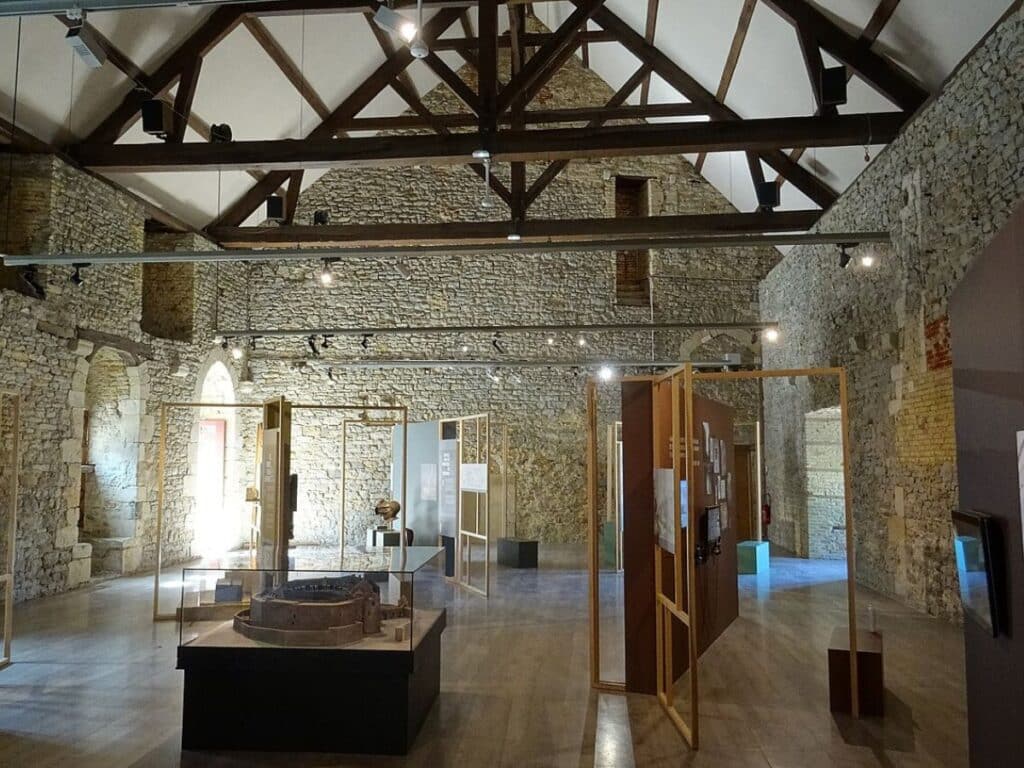Interior of the Château Comtal Museum in Carcassonne, displaying a collection of medieval artifacts within the stone walls of the historic fortress, under a modern wooden beam ceiling.
