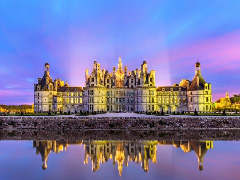 Twilight view of the Château de Chambord in the Loire Valley, a Disney movie location in France, reflecting on the water with a dramatic sky painted in hues of purple and pink, showcasing the castle's distinctive French Renaissance architecture.
