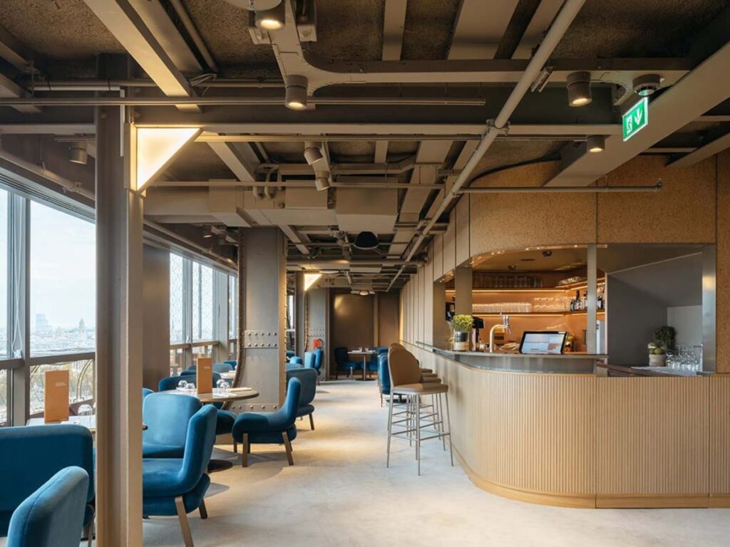 Interior of Madame Brasserie restaurant at the Eiffel Tower with modern decor, featuring blue upholstered chairs, a curved wooden bar counter, and large windows providing a view of the city.