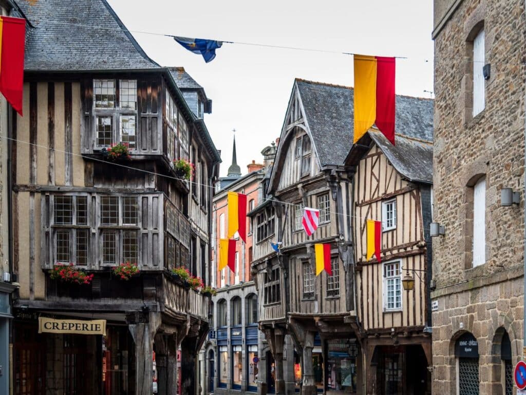 A narrow medieval street in Dinan, Brittany, featuring half-timbered buildings with overhanging upper stories, adorned with flower boxes and colorful flags, capturing the charm of this historic town.