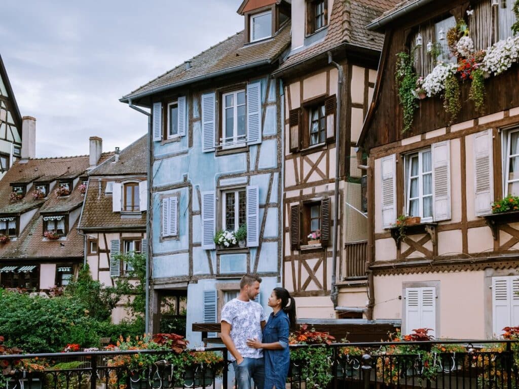 A couple stands close together on a bridge adorned with flowers, with traditional half-timbered houses in the background, reflecting a quaint European village atmosphere. The man gazes at the woman while she looks off to the side, both seemingly enjoying the charming surroundings.