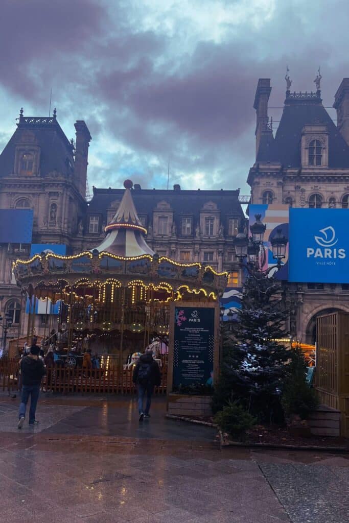 Lit carousel in front of a Parisian city hall under a moody overcast sky.