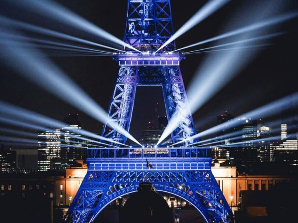 "Night view of the Eiffel Tower illuminated in blue light with radiant beams emanating from the structure, set against a backdrop of the Paris skyline.